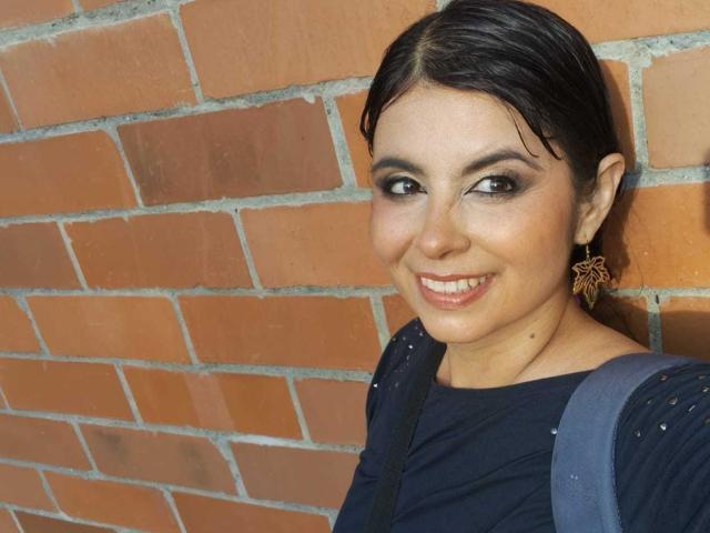 ScarletRebel hat dunkles Haar, einen smokigen Augen-Look und ein Lächeln, bei dem man ihre Zähne sieht. Sie trägt ein dunkles Oberteil mit Sterne-Ohrringen und einem blauen Rucksack. Ihre Brüste sind klein..