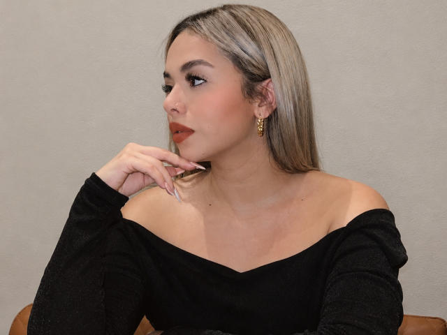Zoe, met blond haar, een zwarte top met open schouders, gouden oorbellen en rode lippenstift, kijkt opzij met haar hand in de buurt van haar mond, waarbij haar lange, puntige nagels te zien zijn..