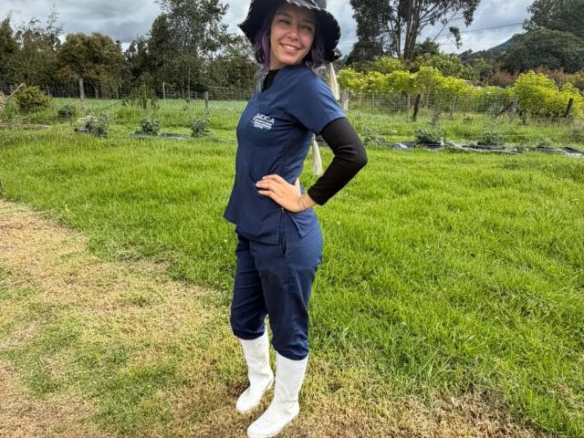 DarckVelvet stands in a grassy field wearing a dark blue uniform, black long-sleeve shirt, white boots, and a wide-brimmed hat, smiling with her hand on her hip.
