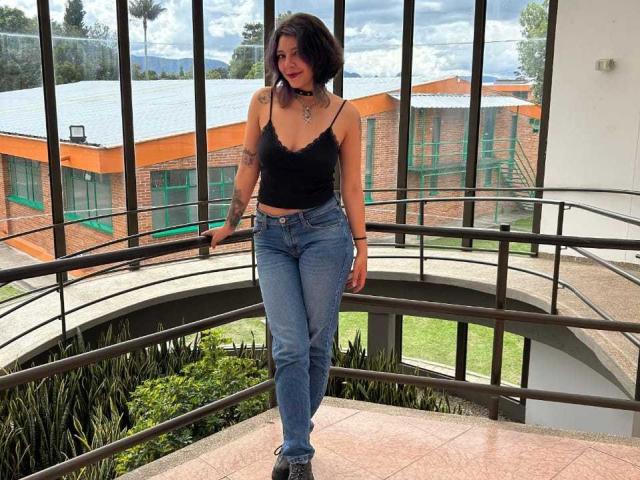 DarckVelvet with short black hair, black lace top, blue jeans, and black shoes, standing on a curved metal balcony, showing her small tits and tattooed arms.