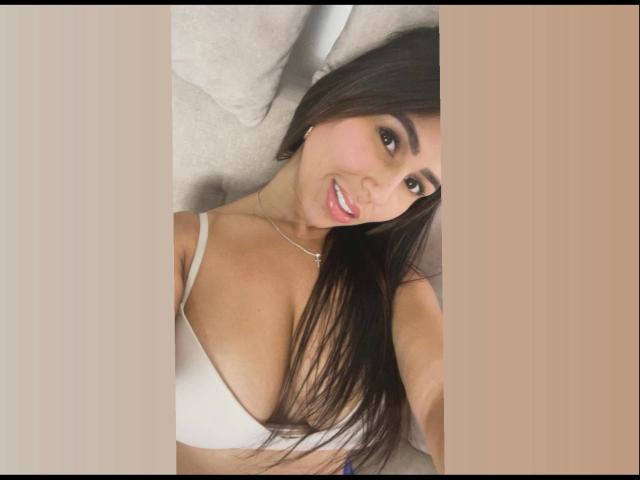 NahomyStone mit langen, braunen Haaren, lächelnd, trägt einen weißen BH, zeigt ihre Brüste, liegt auf einem beigen Sofa und macht ein Selfie. Ihre Brüste sind mittelgroß und sie ist süß..