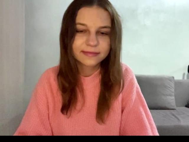 NievesAveles, con cabello castaño, vestida con un suéter rosa, sonriendo ligeramente, mirando hacia abajo, tiene un rostro lindo, pechos pequeños y un tono de piel claro..