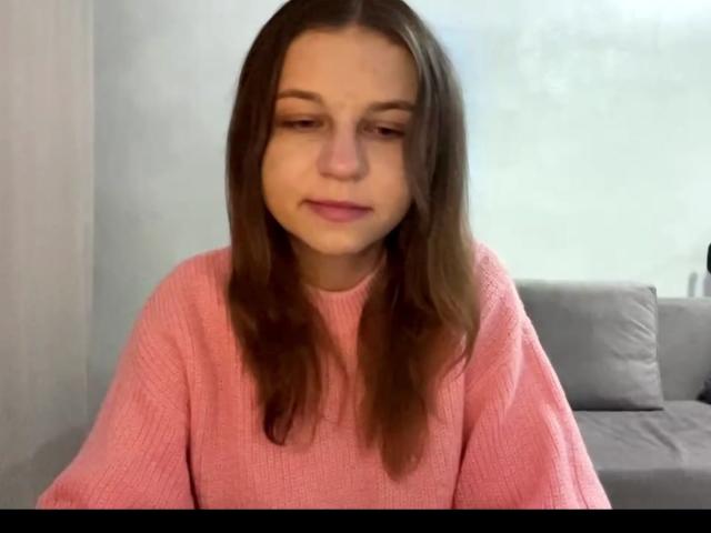 Nieves Aveles, con cabello castaño largo, un suéter rosa y una expresión neutra, mirando ligeramente hacia abajo, tiene senos pequeños y está sentada en una habitación con un sofá gris..
