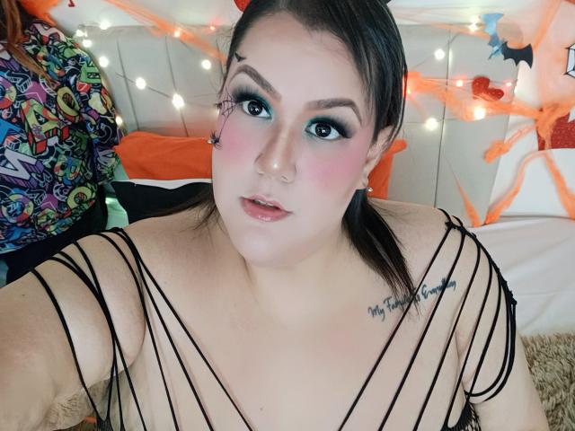 GiiannaMorrys met donkerbruin haar in vlechtjes, draagt een zwart topje met spaghettibandjes, waardoor haar borsten zichtbaar zijn, groene oogschaduw, roze blush en een tatoeage met de tekst "My Tattoo Family" op haar borst..