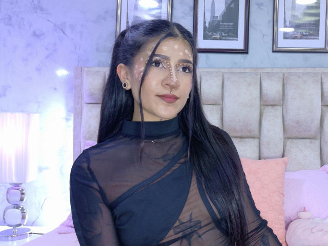 LuciadelaVega with long black hair, sheer black top showing her tits, white face paint, red lipstick, and black earrings, sitting on a bed with pink pillows.