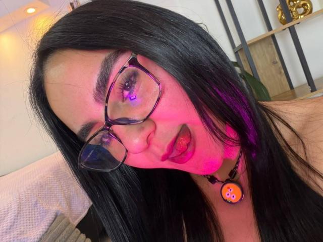 FenixAlthea, avec ses cheveux noirs, ses lunettes et un éclairage rosâtre sur son visage, la langue tirée, portant un collier noir avec un bouton orange rond, dans un autoportrait..