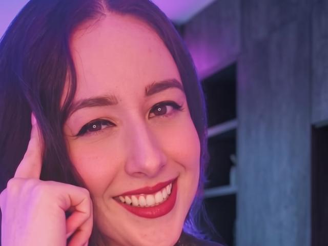 MabyLegrand mit dunkelbraunen Haaren, rotem Lippenstift, lächelnd, die Wange berührend, in violett-blauem Licht, mit braunen Augen, dezentem Make-up und einem süßen, glücklichen Ausdruck..