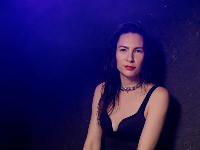 IngaDrein has pale skin, red lipstick, black hair, wearing a black bra and choker, looking at the camera with a slight smile, in a dark, blue-lit setting.