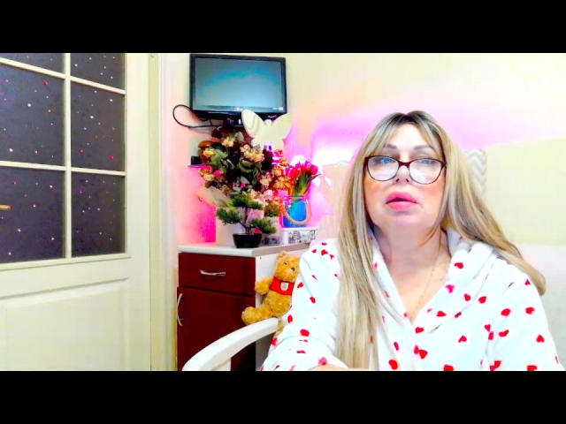 SusieNoreens with glasses, blonde hair, wearing white pajamas with red heart patterns, sitting in a chair, next to a teddy bear and a floral arrangement.