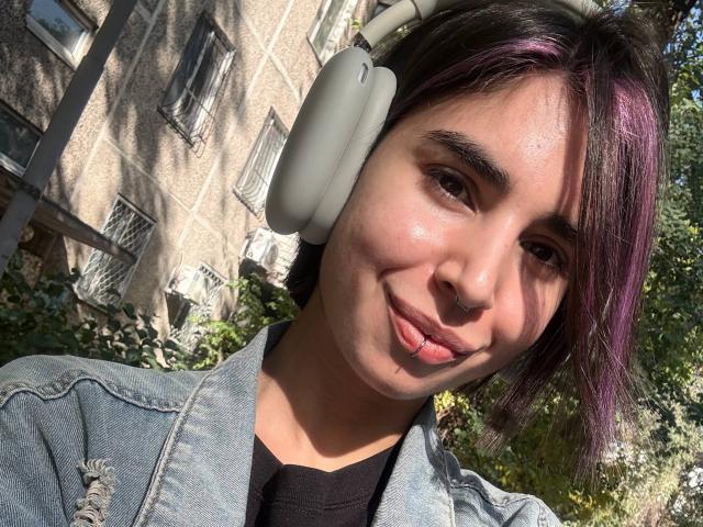 TameikaMacclellan has light brown skin, dark eyebrows, and purple-dyed hair. She's wearing white headphones, a denim jacket, and black shirt, sticking out her tongue.