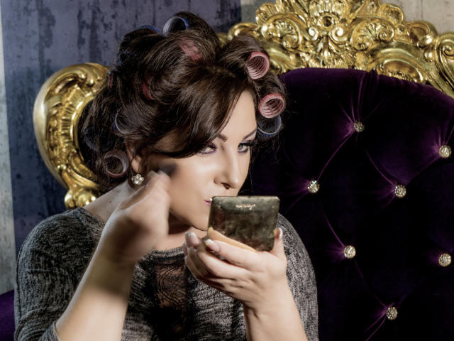 VelvetManttis with curly pink hair, holding a green glass, wearing a grey lace top, looking at a phone, sitting on a purple velvet chair with a gold ornate back.