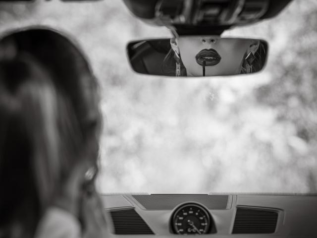 Black-and-white photo showing LucyGrace's reflection in the car mirror with pouty lips, blurred foreground of LucyGrace's wavy hair, and car dashboard with speedometer in the background.