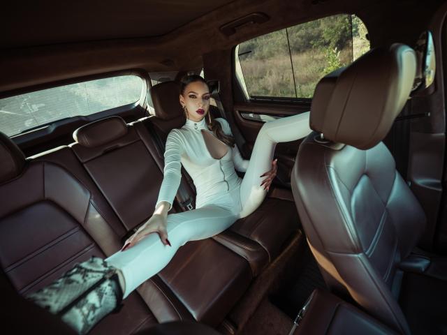 LucyGrace is in a brown leather car, wearing a tight white bodysuit that shows her tits, with her leg up, showing her ass, and looking at the camera.