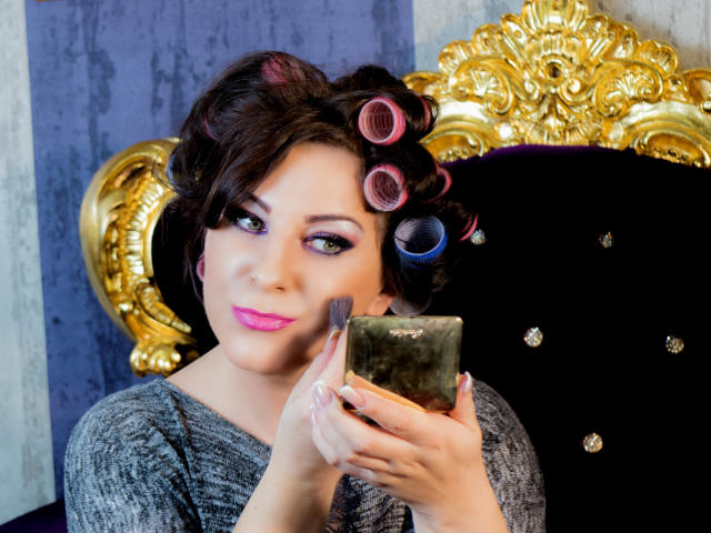 VelvetManttis with curly pink and blue hair rollers, gray sweater, pink lipstick, holding green gemstone, black velvet chair with gold ornate back.