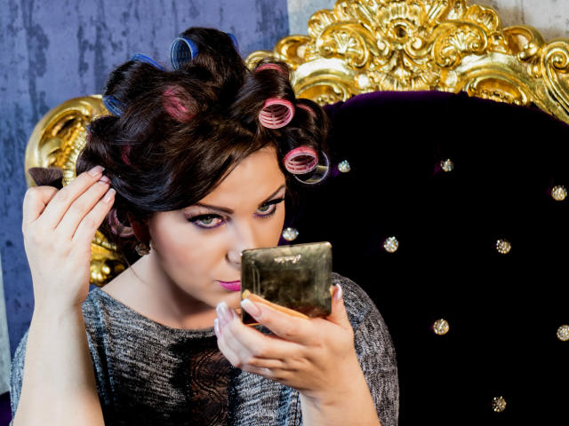 VelvetManttis with curly hair in pink and blue rollers, wearing a gray textured top, holding a gold mirror, looking into it with a slightly annoyed expression.