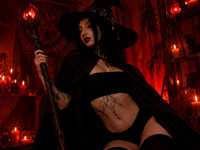 WenMewborn mit schwarzem Hexenhut, schwarzem trägerlosem Oberteil, schwarzer Unterwäsche und Oberschenkelhohen Socken, die einen Stab hält, rot beleuchteter Raum, Tattoo auf ihrem flachen, sexy, freiliegenden Bauch, dunkles Make-up..