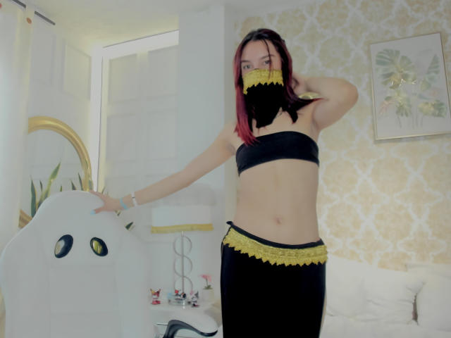 Sahara69 with pink-dyed hair, black and yellow lace mask, black strapless top, and black skirt with yellow lace, standing in a room, hand on white chair.