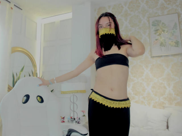 Sahara69 with pink hair, black lace mask, black strapless top, and black lace-trimmed skirt, standing in a room, hand on white chair with cute eyes, showing her ass.