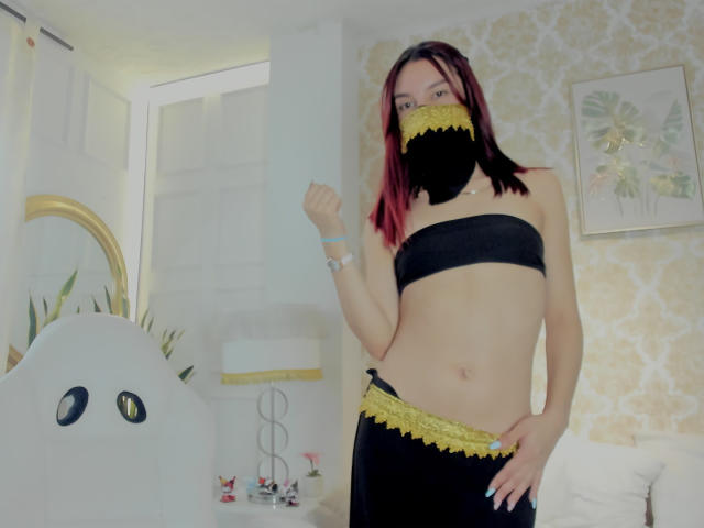 Sahara69 with red-tipped black hair, wearing black lace mask and matching black strapless top and skirt with yellow lace, showing her midriff and fist in a bedroom.