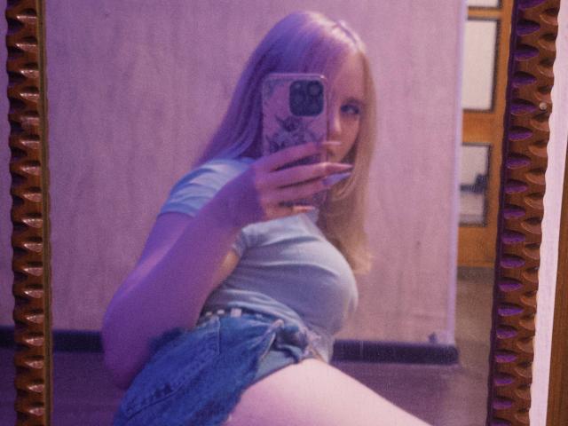TiffaniMacandog, com cabelo loiro, vestindo uma camiseta cinza justa e shorts jeans, tirando uma selfie no espelho, mostrando seus seios e segurando um celular com uma capa rosa..