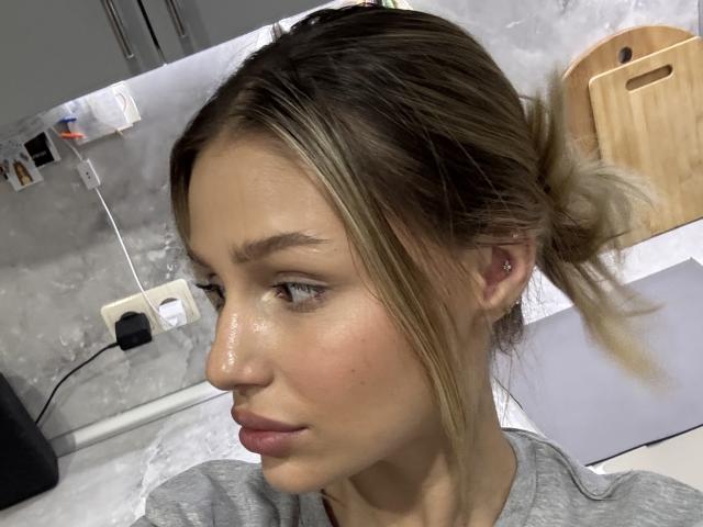 BlackAndWhiyte hat eine glatte Haut, einen blonden Pferdeschwanz, eine kleine Nase, dezentes Make-up, ein graues Hemd und ein unauffälliges Ohrpiercing. Sie blickt zur Seite und hat leicht geöffnete, rosafarbene Lippen..
