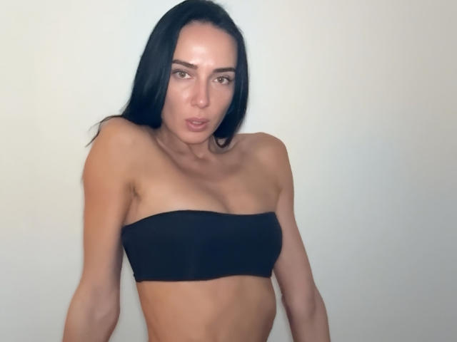 Fridaa with long black hair, wearing a black strapless top, has small tits, a slim body, and a slightly open mouth, standing against a plain white background.