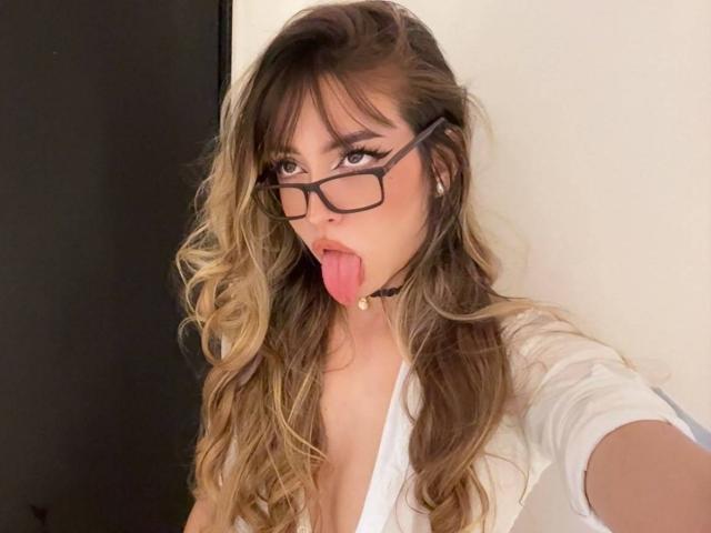 AmeliaCollins with long wavy blonde hair, glasses, and a white shirt, tongue out, wearing a black choker, has a cute face and is looking at the camera.