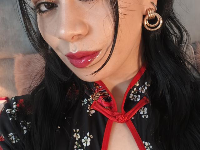 Linda Mancini met zwart haar, rode lippenstift, glanzende lippen, die een zwarte jurk met bloemenprint en rode accenten draagt, en gouden oorringen, close-up, gezicht licht gekanteld. Borsten gedeeltelijk zichtbaar..