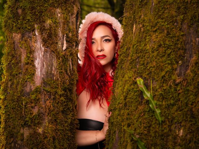 ScarlettthVega with red hair, pink hat, and red lipstick, standing between mossy trees, showing her tits, arms crossed, looking at the camera with a seductive smile.
