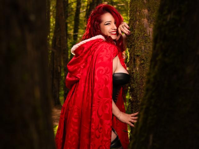 ScarletthVega with red hair, wearing a red ornate cape, black strapless top, standing in a forest, smiling, touching a mossy tree, showing her tits, and red lipstick.