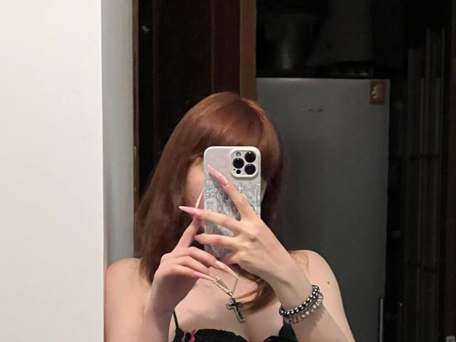 LeomaHaq with red hair, pink nails, black bra, holding phone, covering face, necklace, bracelet, mirror selfie, visible tits, fridge in background.