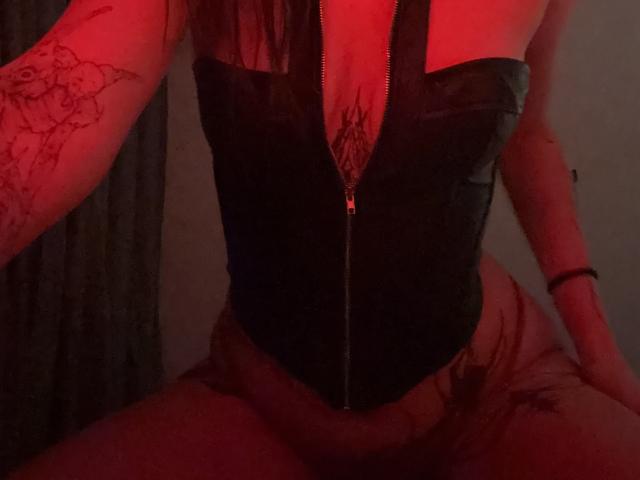 Simonne Brustkern con un corpiño negro con cremallera, iluminación roja, mostrando sus pechos y tatuaje púbico, con un tatuaje floral en el brazo, sentada con las piernas separadas, sin nada puesto debajo..