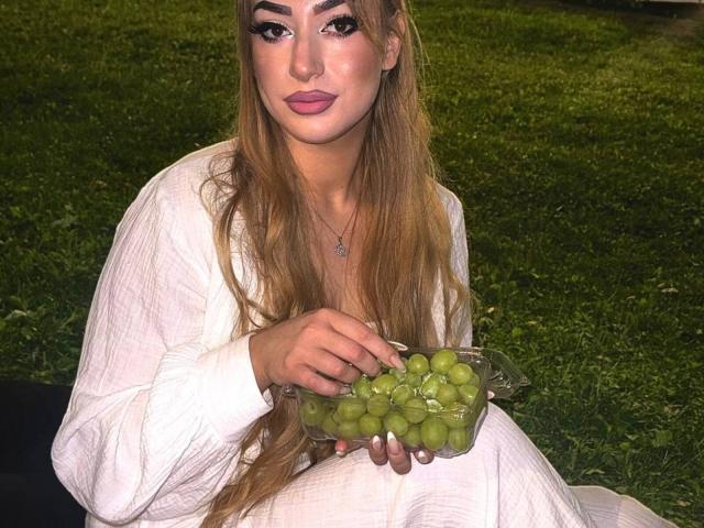 Elvira Crichlow, con cabello rubio largo, vestido blanco, sosteniendo uvas verdes en un recipiente de plástico transparente, sentada sobre la hierba, con labios rosados y delineador de ojos llamativo..