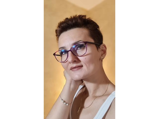 Anne Ross hat kurze, braune Haare, eine violette Brille und ein leichtes Lächeln, wobei ihre Hand auf ihrer Wange liegt. Sie trägt ein weißes Oberteil und eine goldene Halskette..