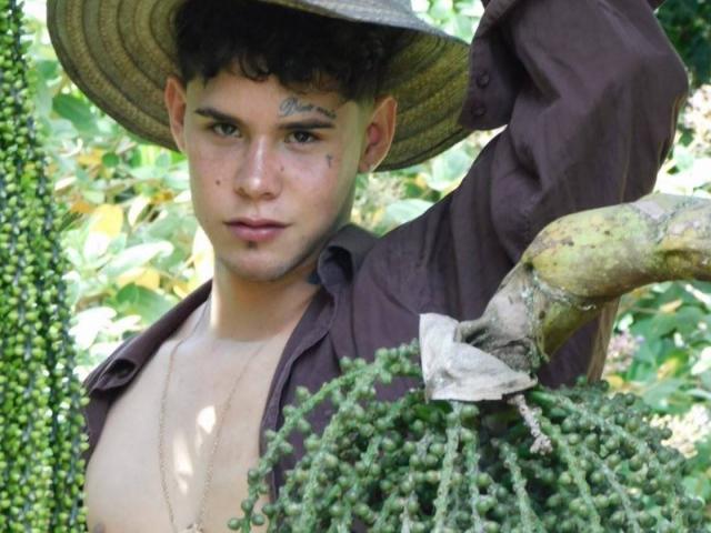 DerrickBigX with curly brown hair, freckles, and a nose ring, shirtless, wearing a brown hat, holding a tree branch with green clusters, looking at the camera.