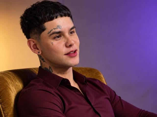DerrickBigX with short black hair, tattoos on forehead and neck, dark maroon shirt, ear piercings, sitting on golden chair, smiling, purple and yellow gradient background.