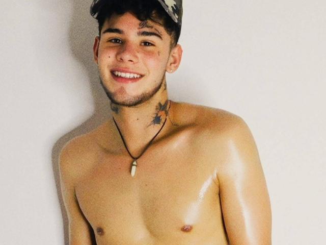 Shiny DerrickBigX with a cute smile, dark hair, and a small beard, wearing a cap, necklace with a white stone, and star tattoos on his neck. Shirtless, glistening skin.
