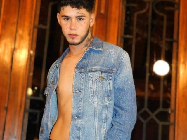DerrickBigX, shirtless with a blue denim jacket, has tattoos on his neck and upper chest. He's standing with arms behind his back, looking slightly to the side.