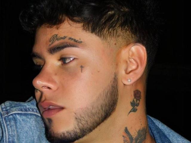 DerrickBigX has curly dark hair, a nose ring, and tattoos including "most me" on his forehead, a cross on his cheek, and a rose on his neck.
