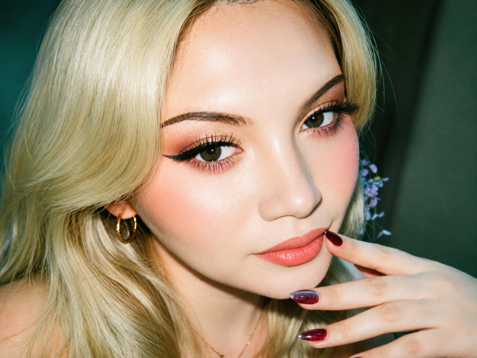 ScarletteCathy with blonde hair, pinkish-orange eyeshadow, red lipstick, and maroon nails, touching her chin, wearing gold hoop earrings, looking directly at the camera.