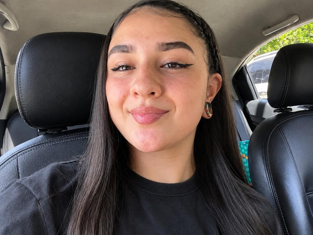 SamyBennet has a cute smile with pink lips, black eyeliner, and long straight brown hair, wearing a black shirt and hoop earrings, sitting in a car's leather seat.
