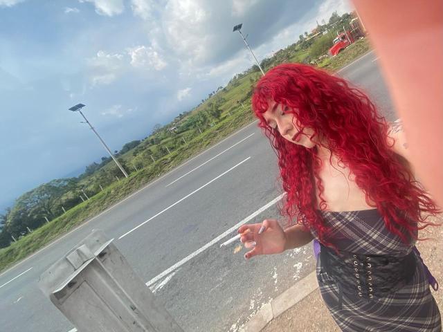 GaiaDoll with red curly hair, strapless plaid dress, and visible tits, standing on roadside, holding a cigarette, overcast sky, and empty road in background.