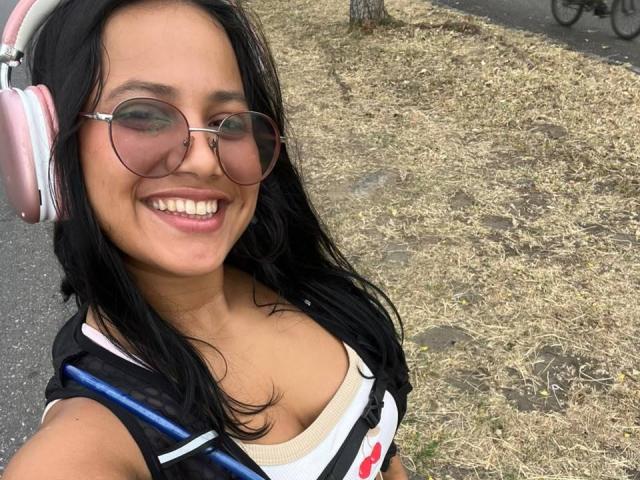 AlanaLuxe mit schwarzen Haaren, die rosa Kopfhörer trägt, runde Sonnenbrille, lächelnd, zeigt kleine Brüste in einem schwarz-weißen Oberteil, steht auf einem grasbewachsenen Bürgersteig..