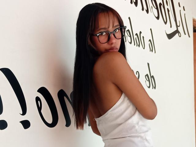 LovelyConnie with long black hair, glasses, and a white towel covering her ass, looking back over her shoulder, showing her bare back and a slight smile.
