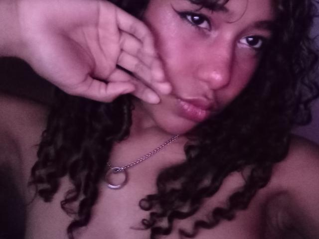 LovelyConnie with curly dark hair, pouty lips, and a silver necklace with a circular pendant, covering her nose with her hand. Her bare shoulders are visible.
