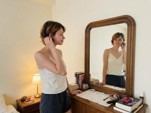 Charlsie Lunning, avec ses courts cheveux bruns, son débardeur blanc et son short noir, se tient devant un miroir, touchant son oreille. On peut voir son reflet. Un commode en bois est visible, avec du maquillage et une lampe..