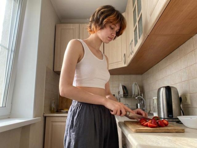Charlsie Lunning, avec ses courts cheveux bruns, son débardeur blanc et sa jupe grise, coupe des tomates rouges sur une planche à découper dans une cuisine baignée de soleil. Elle a de petites seins et une silhouette mince..