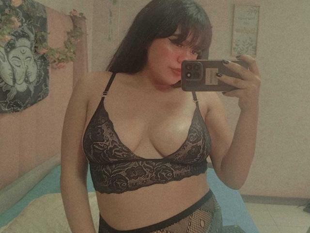 NicoleSamanta with dark hair, wearing black lace bra showing her tits and fishnet pants, taking a mirror selfie with a phone, pink blush on her cheeks.