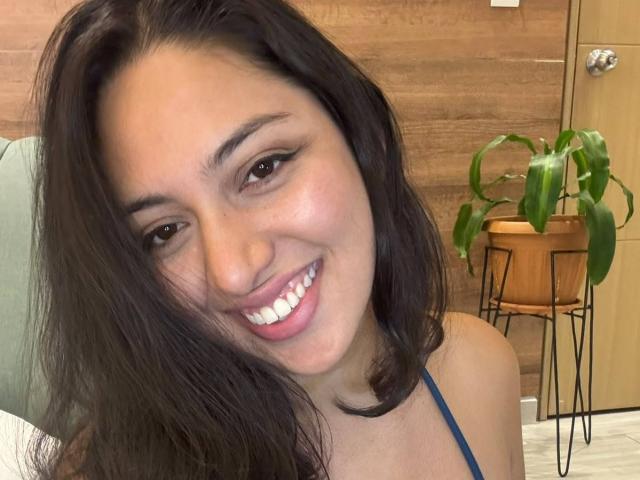 VictoriaLusty has dark, wavy hair, brown eyes, and a bright smile showing white teeth. She's wearing a blue top, and her ass is partially visible.