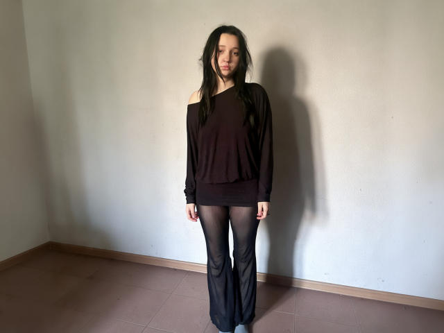 ElleyMolly has long black hair, a slim build, and wears a black off-shoulder top with sheer black pants, standing with a shadow on a plain wall.